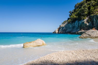 Pürüzsüz beyaz çakıl taşları, Sardinya 'nın Cala Goloritze sahilinde yükselen güneşli kireçtaşı kayalıkları ve yemyeşil yeşil kayalıklarıyla el değmemiş bir plajda hafif turkuaz dalgalarla karşılaşıyor. Açık mavi gökyüzü ve canlı su sakin bir yaz atmosferi yaratır..