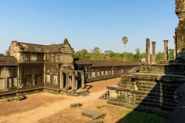 Öğleden sonra güneş ışığı, Angkor Varilleri 'nin yıpranmış kumtaşı galerisini ve süslü giriş köşkünü vurguluyor. Kuru çimenler ve yapayalnız bir palmiye ağacı tarihi Khmer mimarisini Siem Reap 'te berrak bir mavi gökyüzüne karşı çerçeveler.