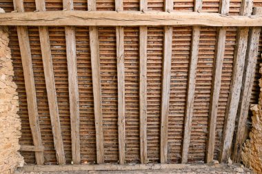 Yıpranmış ahşap kirişler ve sıra sıra kil kiremitler geleneksel bir kırsal çatının içini oluşturuyor. Yapıda doğal dokular, düzensiz çizgiler ve tarihi yapı işçiliği yer alıyor..