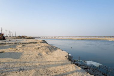 Geniş kumlu nehir kıyısı Hindistan 'ın Prayagraj kentinde sakin Ganj Nehri ile birleşiyor ve ufuk boyunca uzanan uzun bir köprü berrak mavi gökyüzünün altında. Nadir yapılar ve elektrik direkleri sakin manzaraya ince detaylar ekler..