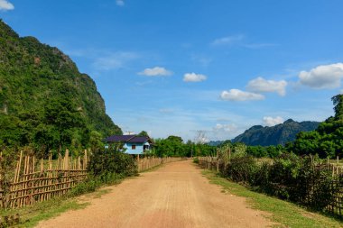 Bambu çitleriyle çevrili toprak bir yol Laos kırsalındaki yemyeşil ve dramatik kireçtaşı tepelerinin ortasındaki mavi ahşap bir evin yanından geçiyor. Parlak güneş ışığı ve dağınık bulutlar huzurlu ve açık manzarayı güçlendirir..