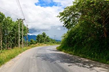 Sessiz bir asfalt yol, verimli bitki örtüsü ve elektrik direkleriyle çevrili genç ağaçların arasından, kuzey Laos 'ta parçalı bulutlu bir gökyüzünün altında uzak mavi tepeler boyunca esiyor. Sahne, kırsal bir yolun sakin atmosferini yakalıyor.