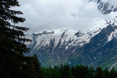 Yoğun yeşil bir ormanın üzerinde kar lekeli dağ yamaçları yükseliyor. Mont Blanc 'taki engebeli zirveleri kısmen gizleyen dramatik bulutlar var..
