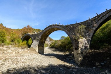Yunanistan 'ın Epirus kenti Kipoi yakınlarındaki renkli sonbahar yaprakları ve engebeli tepelerle çevrili üç zarif taş kemer ile tarihi Plakidas Köprüsü kuru, kayalık bir nehir yatağını kaplar. Parlak gün ışığı desenli duvarcılığı ve doğal manzarayı gözler önüne seriyor.
