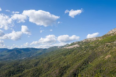 İtalya, Sardunya 'da Baunei yakınlarında, tüylü beyaz bulutlarla kaplı, parlak mavi bir gökyüzünün altında yoğun yeşilliklerle kaplı yuvarlanan tepeler. Güneş ışığı Akdeniz 'in engebeli hatlarını ve doğal dokularını vurgular.