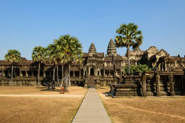 Düz bir taş yol, Angkor Vat 'ın onur terasına uzanır. Uzun palmiye ağaçları ve tarihi kumtaşı kuleleri tarafından çerçevelenir. Yıpranmış Khmer mimarisi açık mavi gökyüzünün altında duruyor ve UNESCO 'nun anıtsallığını vurguluyor.