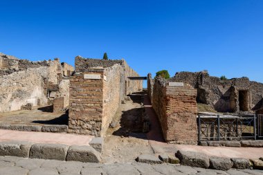 Tarihsel tuğla ve volkanik taş duvarlar İtalya 'nın Pompeii kentindeki arkeolojik alanda dar bir giriş oluştururlar. Sahnede sert taş ustaları, güneş ışığı dokuları ve antik Roma kalıntılarının üzerinde canlı bir mavi gökyüzü yer alıyor..