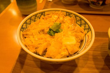 Geleneksel bir Japon oyakodonu taze tavuk ve kabarık yumurta seramik bir kasede pilav üzerine servis edilir. Sıcak ortam aydınlatması parlak dokuları ve rahatlatıcı sunumu vurgular.