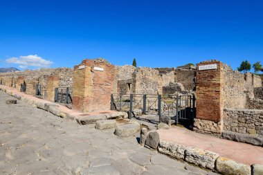 Bir dizi yıpranmış tuğla ve taş girişi İtalya 'nın Pompeii kentindeki tarihi Roma caddesinde yer alıyor. Sahne desenli duvar ustalığı, demir parmaklıklar ve canlı mavi bir gökyüzü içeriyor, bu antik şehir planının korunmuş halini vurguluyor.