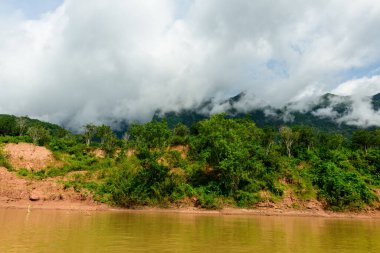 Yoğun bulutlar, kuzey Laos 'taki Muang Ngoi yakınlarındaki Nam Ou nehri boyunca yemyeşil bir tepenin üzerinde sürükleniyor. Sahnede canlı bitki örtüsü, aşınmış nehir kıyıları ve tropikal manzaraya çarpıcı bir ışık yer alıyor..