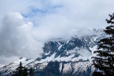 Aiguille de Blaitiere 'in dik kayalık yamaçları kalın bulutlarla kısmen örtülmüş. Fransız Alpleri' ndeki dramatik alp manzarasını çevreleyen sürekli yağan kar ve yeşil ağaçlarla kaplı..