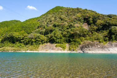 Canlı turkuaz su, parlak güneş ışığıyla aydınlatılan ve berrak mavi bir gökyüzü tarafından çerçevelenen, kayalık çıkıntılı, ağaçlarla kaplı bir yamaçtan nazikçe geçer..