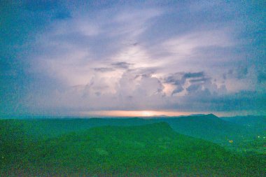 Tek bir yıldırım, engin gök gürültüsü bulutlarının altında ufku aydınlatır. Uzak tepeler, kasvetli mavi bir alacakaranlık gökyüzüne karşı siluetlenir. Sahne dramatik bulut oluşumlarını, ince eğimleri ve atmosferik gerilimi yakalar.