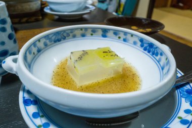 Şeffaf kivi jölesiyle kaplanmış hassas bir tofu karesi sığ bir altın sos havuzunda, dekoratif mavi ve beyaz bir porselen kasede servis edilir. Kompozisyon pürüzsüz dokuları ve rafine Japon yemek atmosferini vurguluyor..