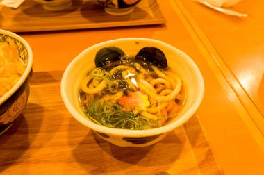 Buharda pişen udon erişte çorbası kalın erişte, dilimlenmiş yeşil soğan, yosun ve şeffaf et suyunda dekoratif sebze dilimi içerir. Sıcak, altın ışıklandırma parlak yüzeyi vurgular ve ahşap bir tepside sunum yapar..