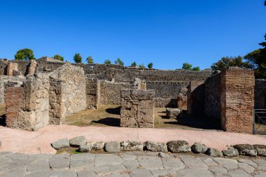 Yıpranmış volkanik taş ve tuğla duvarlar İtalya 'nın Pompeii kentindeki arkeoloji sahasında canlı bir mavi gökyüzünün altında duruyor. Sahne, pürüzlü duvarcılık, açık alanlar ve bu antik Roma şehrinin tarihi dokularını vurguluyor..