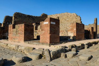 İtalya 'nın Pompeii arkeoloji sahasında güneş ışığıyla aydınlanan bir cadde boyunca eskimiş tuğla sütunlar ve volkanik taş kalıntıları duruyor. Sahne, duvarları, dokulu yüzeyleri ve açık mavi gökyüzünün altındaki tarihi şehir yapısını ön plana çıkarıyor..