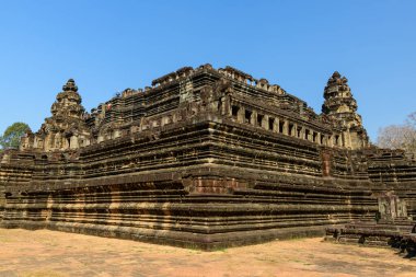 The monumental Baphuon temple features stepped sandstone terraces and intricate Khmer architecture under a clear blue sky in Angkor, Cambodia. Warm sunlight highlights the weathered stonework and geometric lines of this historic site.