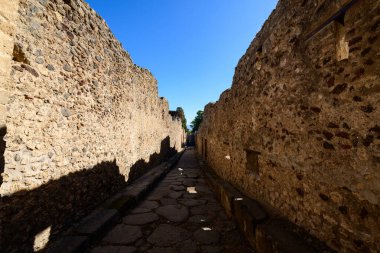 Pompei 'nin arkeolojik bölgesindeki canlı mavi gökyüzünün altında yıpranmış taş duvarlarla çevrili antik bir kaldırım taşı caddesi. Sahnede desenli duvarcılık, keskin gölgeler ve bu Roma şehrinin tarihi şehir düzeni yer alıyor..