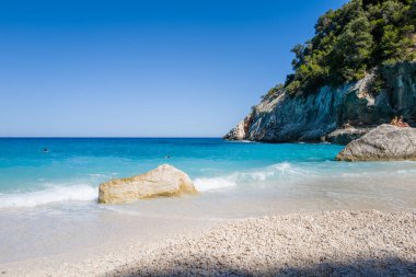 Beyaz çakıl taşları ve büyük bir taş el değmemiş bir sahilde turkuaz dalgaların kenarında oturur. Sardunya 'nın Cala Goloritze şehrinde bulutsuz mavi bir gökyüzünün altında dramatik kireçtaşı uçurumları ve yemyeşil bir gökyüzü yükselir..