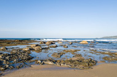 Kotohikihama Sahili 'nde berrak mavi bir gökyüzü altında sığ havuzları ve yumuşak dalgaları olan kayalık gelgit düzlükleri geniş bir manzara oluşturur..