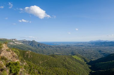 Güneşli yeşil dağ yamaçları, Sardunya 'nın Baunei ve Dorgali yakınlarında ufukta görünen Akdeniz' le birlikte geniş bir kıyı ovasına doğru iniyor. Açık gökyüzü ve dağınık bulutlar parlak, geniş bir atmosfer yaratır..