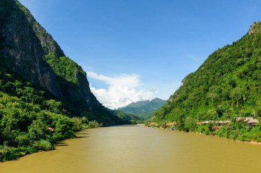 Laos 'un Nong Khiaw kentindeki Nam Ou nehrinin her iki yakasındaki sarp yeşil kireçtaşı kayalıkları açık mavi gökyüzünün altında yükseliyor. Geleneksel nehir kenarındaki evler ve yoğun tropikal orman kuzey Laos 'ta dramatik bir doğal manzara yaratır..