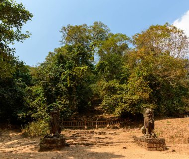 Kamboçya 'daki Siem Reap yakınlarındaki Phnom Bakheng üssündeki yoğun ormana çıkan kırsal bir merdivenden iki antik taş aslan muhafızı tarafından kuşatıldı. Sahnede kuru toprak, yapraklı ağaçlar ve açık mavi gökyüzünün altında güneşli bir açıklık yer alıyor..