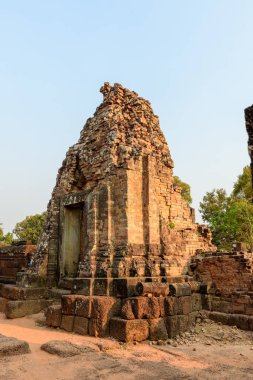 Kamboçya 'daki Pre Rup tapınağındaki eski bir kırmızı tuğla tapınak kulesinin güneşli manzarasında aşınmış taş bloklar ve aşınmış taş bloklar yer alıyor. Tarihi yapı kumlu zemin ve dağınık yeşillikler arasında duruyor. Angkor 'un mirasını yansıtıyor.