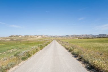 Açık mavi gökyüzünün altından İspanya 'daki Bardenas Reales' in engebeli tepelerine uzanan uzun toprak bir yol geçmektedir. Manzara güneşli, kuru ve geniş, özgürlük ve macera hissi uyandırıyor..