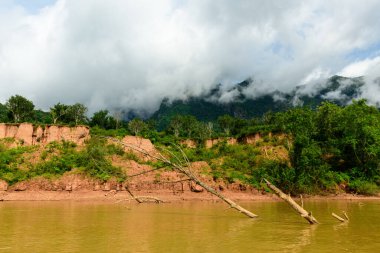 Laos 'un kuzeyinde Muang Ngoi yakınlarında Nam Ou nehri boyunca alçak bulutlar süzülüyor. Erotik kayalıklar, düşen ağaç gövdeleri ve yoğun yeşil bitki örtüsü dramatik bir gökyüzünün altında dokulu bir manzara yaratır..