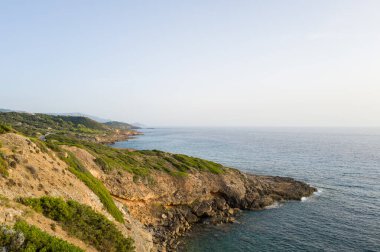 Güneşli kayalık kayalıklar ve yeşil Akdeniz ovaları, Sardinya 'nın Alghero ve Bosa kıyıları boyunca uzanan sakin mavi denizi gözler önüne serer. Manzara, toprak dokuları, yumuşak yamaçlar ve berrak bir gökyüzünün altında sakin bir atmosfere sahiptir..