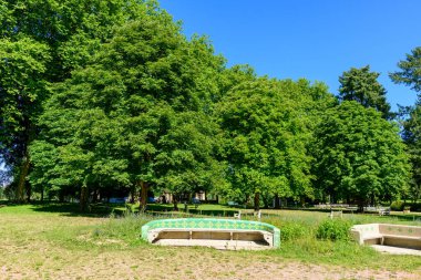 Ornate concrete benches with green tile accents sit on a grassy lawn, surrounded by dense leafy trees and vibrant greenery under a clear blue sky in Saint-Honore-les-Bains.