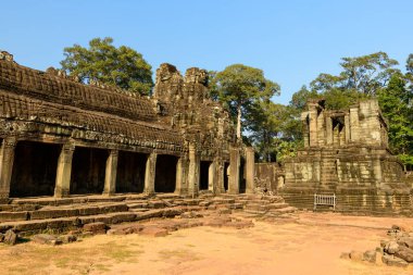 Öğleden sonra güneş ışığı, sütunlu galeriye sıcak bir tonla vuruyor ve Angkor, Kamboçya 'daki Bayon Tapınağı' nın kulesine basıyor. Yıpranmış kumtaşı yapılar, açık mavi gökyüzünün altındaki orman ağaçlarının arasında duruyor, tarihi Khmer mimarisini ve