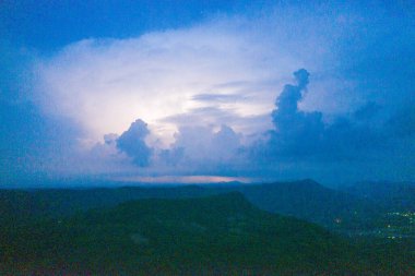 Karanlık bir dağ sırtı üzerinde alacakaranlıkta yükselen yıldırımlarla aydınlanan yüksek fırtına bulutları. Sahnede dramatik bulut oluşumları, katmanlı siluetler ve kasvetli bir alacakaranlık gökyüzü yer alıyor..