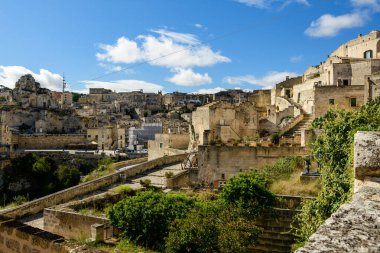 İtalya 'nın Matera kentindeki tarihi taş binaların ve teraslı çatıların geniş bir manzarası, dağınık bulutlu açık mavi bir gökyüzünün altında. Güneş ışığı, antik duvarlar boyunca kireçtaşı mimarisi ve yemyeşil alanları vurguluyor.