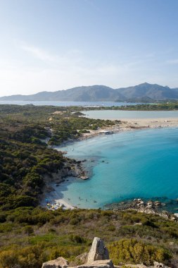 Yeşil tepeler ve Porto Giunco 'daki kumlu plajlarla çevrili turkuaz bir körfez manzarası. Uzak dağlar ve berrak bir gökyüzünün altında sakin bir lagün..