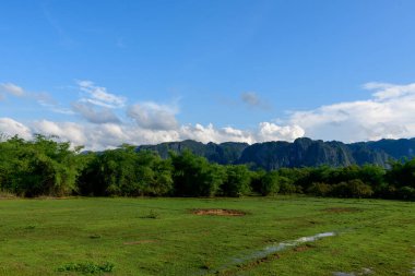 Yoğun yeşil çalılarla çevrili açık çimenli bir alan. Arka planda dramatik kireçtaşı tepeleri yükseliyor. Canlı mavi bir gökyüzü ve dağınık bulutlar altında. Manzarada verimli bitki örtüsü ve Güneydoğu Asya kırsalının tipik bir açık alan hissi yer alıyor.