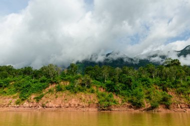 Kuzey Laos 'taki çamurlu bir nehir kıyısındaki yemyeşil orman boyunca alçak bulutlar hareket ediyor. Sahnede yoğun bitki örtüsü, aşınmış toprak ve parlak bir gökyüzünün altındaki sisli dağ yamaçları yer alıyor..