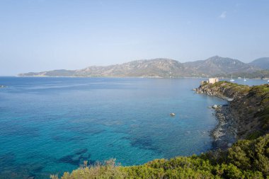Turkuaz berrak sular Cala Caterina 'nın kayalık kıyı şeridi boyunca uzanır. Buruna tünemiş eski bir taş gözlem kulesi ve parlak bir gökyüzünün altında yükselen engebeli dağlar..