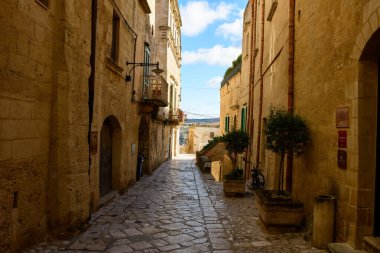 İtalya 'nın Matera kentindeki yıpranmış taş binalar arasında sakin bir kaldırım taşı sokak rüzgarı, sıcak güneş ışığı ve saksı yeşilliği ile tarihi mekana cazibe katıyor..