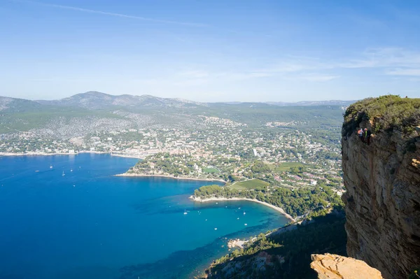 Seyrek bitki örtüsüne sahip kayalık uçurum turkuaz körfez ve kıyı kasabası Cassis 'e bakıyor ve arka planda yuvarlanan tepeler ve açık mavi gökyüzü bulunuyor. Parlak güneş ışığı, engebeli kaya yüzeyi ile sakin Akdeniz arasındaki zıtlığı vurguluyor