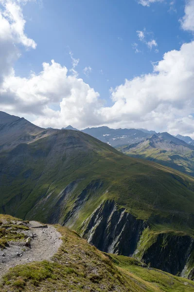 Dar dağ patikaları dramatik uçurumların üzerindeki çimenli bir tepe boyunca esiyor. Güneşli yamaçlar ve engebeli tepeler Val dAoste Alplerinde parlak bir gökyüzünün altında..