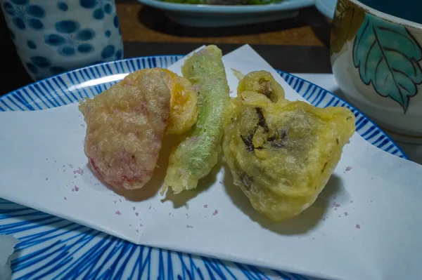 Hafif hırpalanmış üç sebze parçası, zarif Japon sofra takımlarıyla çevrili mavi desenli seramik bir tabağın üzerinde beyaz bir kağıda dizilmiştir. Yakın plan, canlı dokuları, ince renkleri ve ince bir yemek atmosferini ön plana çıkarıyor..