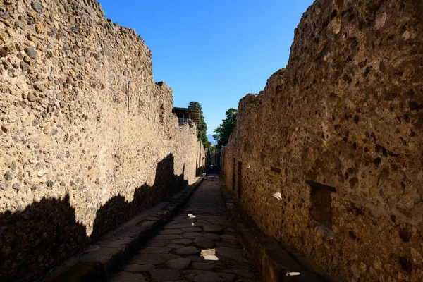 Pompeii arkeolojik alanında aşınmış taş duvarlarla çevrili dar bir cadde, berrak mavi gökyüzünün altında parlak güneş ışığıyla aydınlatılıyor. Sahne desenli duvar ustalığını, derin gölgeleri ve bu Roma kentinin tarihi yerleşimini vurguluyor.