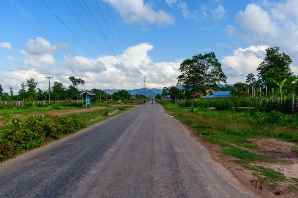 Yeşil tarlalar ve dağınık köy evleri arasında verimli bitki örtüsü ve yüksek ağaçlarla çevrili asfalt bir yol uzanıyor. Tepeler, elektrik hatları ve dramatik bulutlar gökyüzünü doldurarak Laos 'ta sakin bir kırsal bölge yaratıyor..
