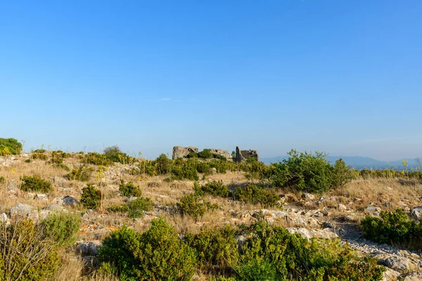 Şatonun antik taş kalıntıları, ufuktaki uzak dağlarla birlikte, kuru çimenler ve alçak çalılarla dağılmış, güneşli, kayalık bir yamacın tepesinde oturur..