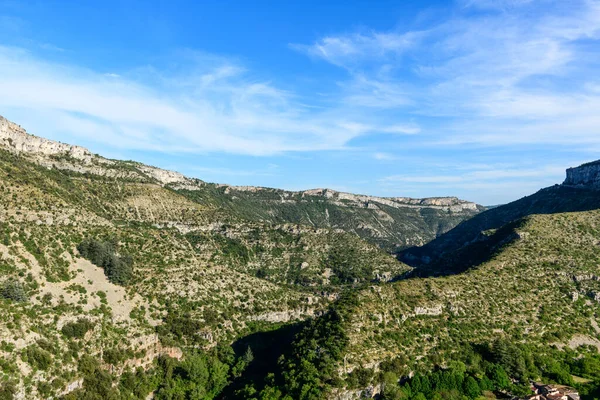 Geniş kireçtaşından tepeler ve engebeli yamaçlar Navacelles Sirki boyunca parlak mavi bir gökyüzünün altında uzanıyor. Parlak güneş ışığı kayalık dokuları güçlendirir ve seyrek yeşil bitki örtüsü geniş bir açık alan hissi yaratır..
