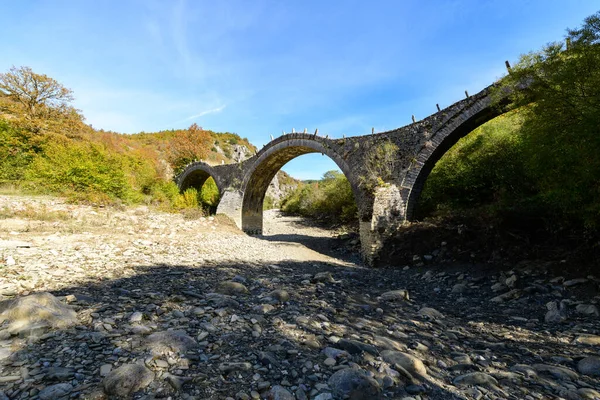 Üç kemerli Plakidas Köprüsü, Yunanistan 'ın Epirus kentindeki Kipoi yakınlarında sonbahar yaprakları ve engebeli tepelerle çevrili kayalık, kuru bir nehir yatağını kaplar. Parlak gün ışığı desenli taş işçiliğini ve Pindus Dağları 'nın doğal manzarasını gözler önüne seriyor..