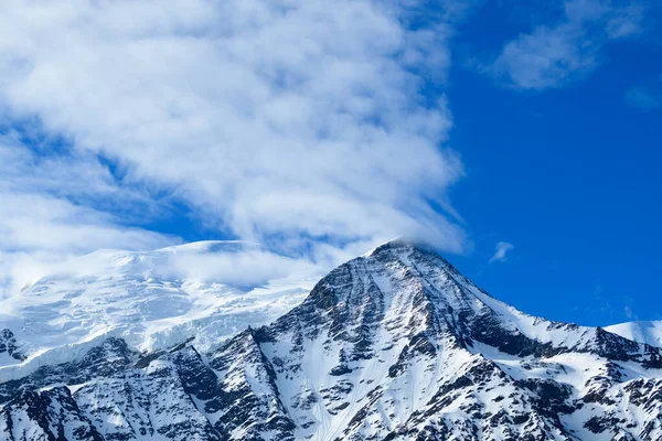 Mont Blanc ve Aiguille du Gouter 'ın keskin zirvesi engebeli karlı yamaçların üzerinde yükseliyor, parlak bir alp atmosferi için canlı mavi gökyüzünde dönen dönen bulutlarla..